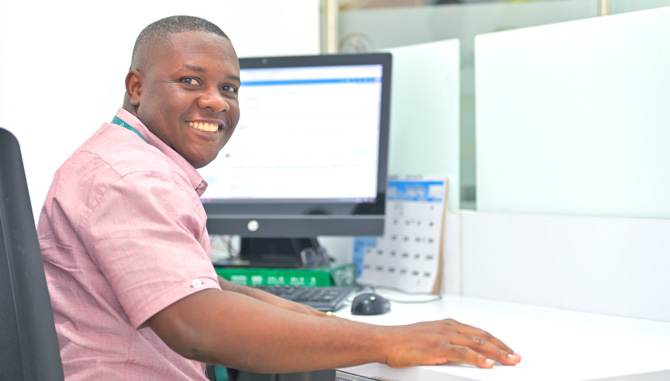 Hombre sentado en una silla sonriendo. En el fondo se muestra la pantalla de un computador.