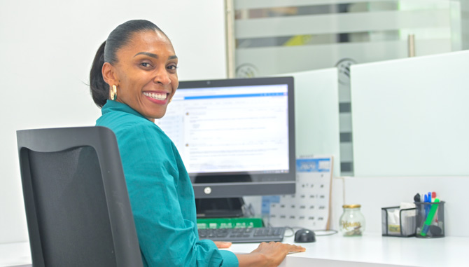 Mujer sentada en una silla sonriendo. En el fondo se muestra la pantalla de un computador.