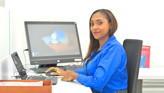 Mujer sentada en una silla sonriendo. En el fondo se muestra la pantalla de un computador.