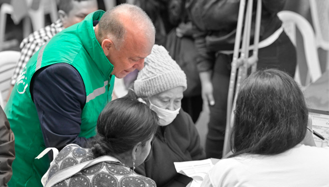 Hombre sentado en una silla sonriendo. En el fondo se muestra la pantalla de un computador.