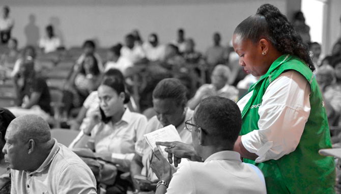 Mujer sentada en una silla sonriendo. En el fondo se muestra la pantalla de un computador.